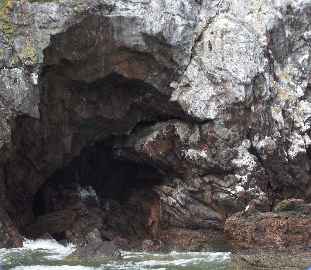 A photo of a dark sea cave, white stained rocks with gentle foam topped waves.